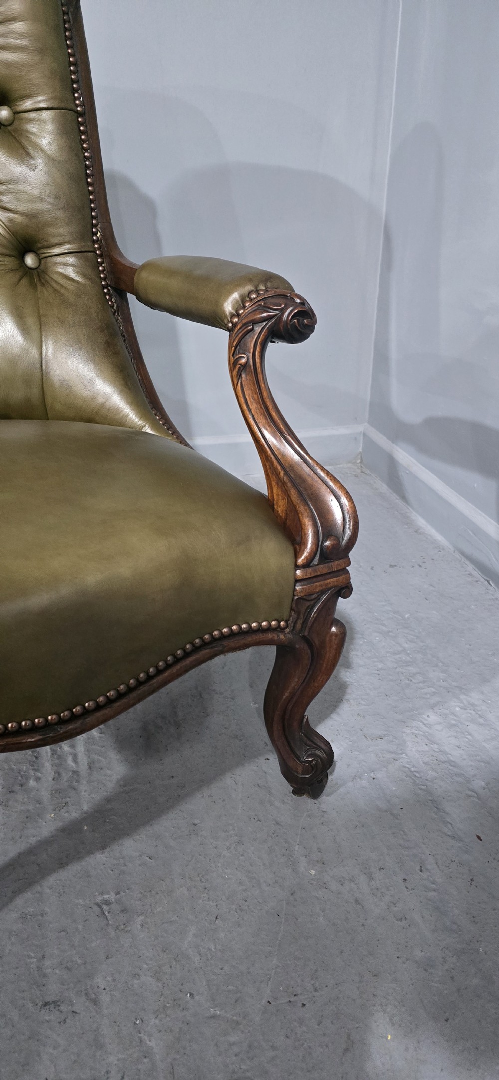 An antique Victorian green leather library chair with deep-buttoned upholstery, carved mahogany frame, scrolled arms, and cabriole front legs on brass castors. View X