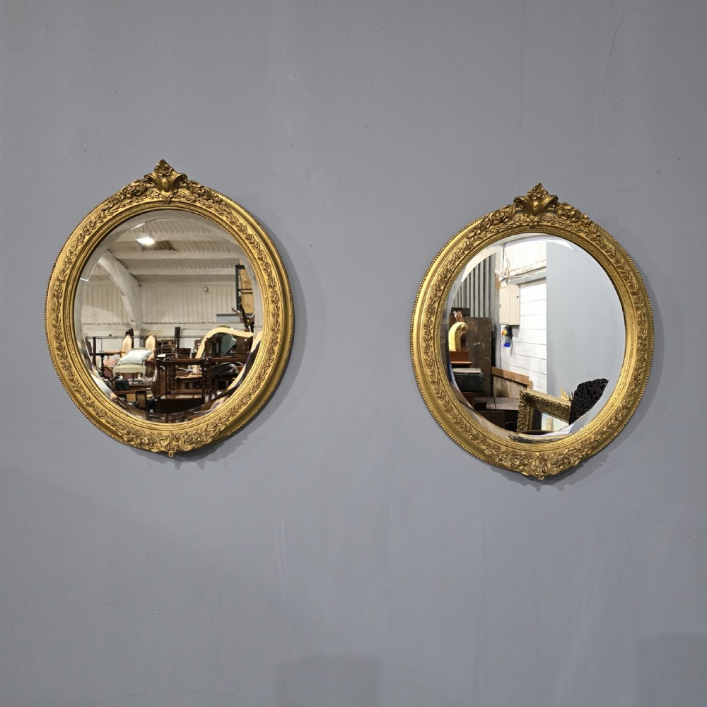 A matching pair of oval giltwood wall mirrors featuring ornate carved top crests and decorative inner rims.