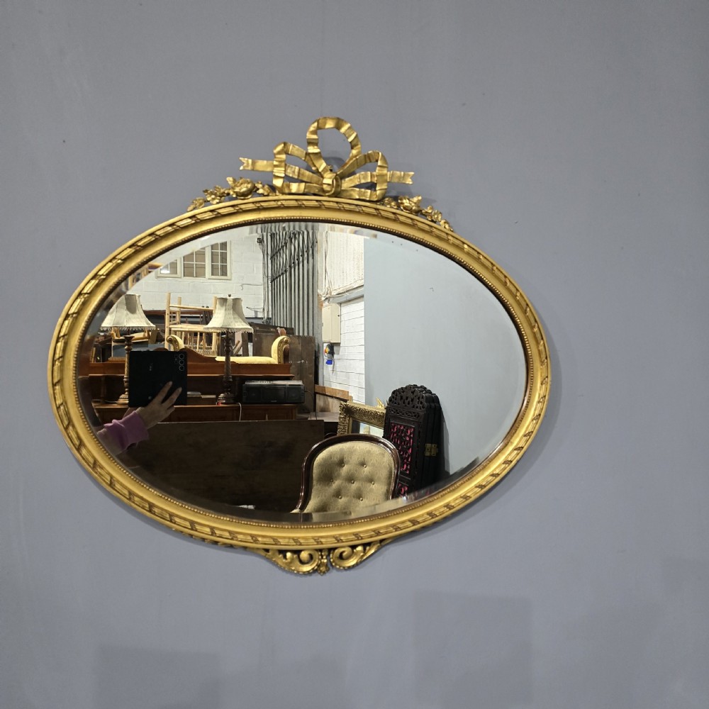 An oval giltwood wall mirror featuring an intricate ribbon bow crest and beaded border decoration.