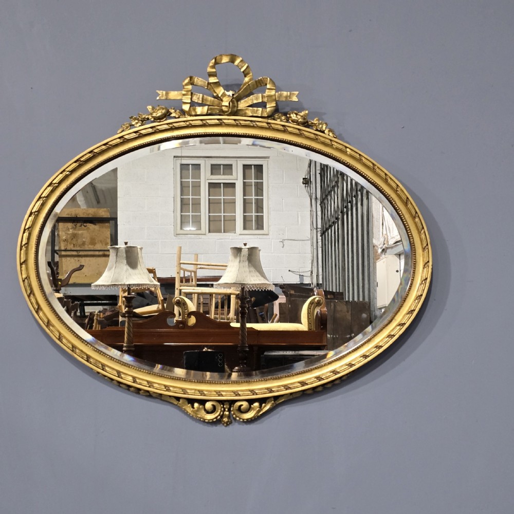 An oval giltwood wall mirror featuring an intricate ribbon bow crest and beaded border decoration. View II