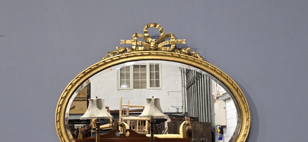 An oval giltwood wall mirror featuring an intricate ribbon bow crest and beaded border decoration. View III