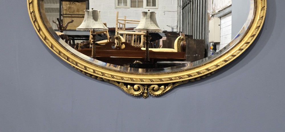 An oval giltwood wall mirror featuring an intricate ribbon bow crest and beaded border decoration. View IV