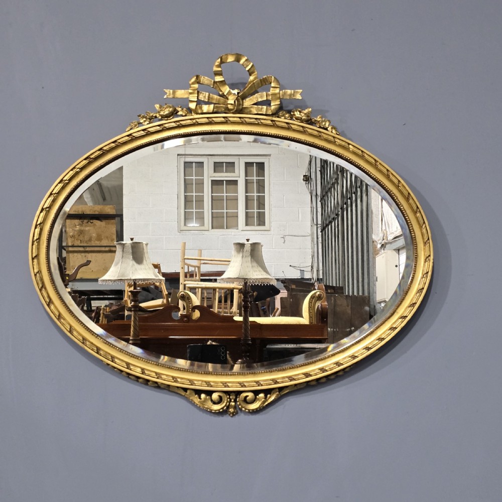 An oval giltwood wall mirror featuring an intricate ribbon bow crest and beaded border decoration. View V
