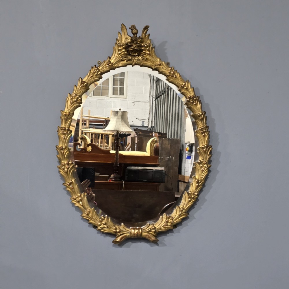 An oval wall mirror featuring a gilded carved wood frame adorned with acanthus leaves and a floral crest top motif.