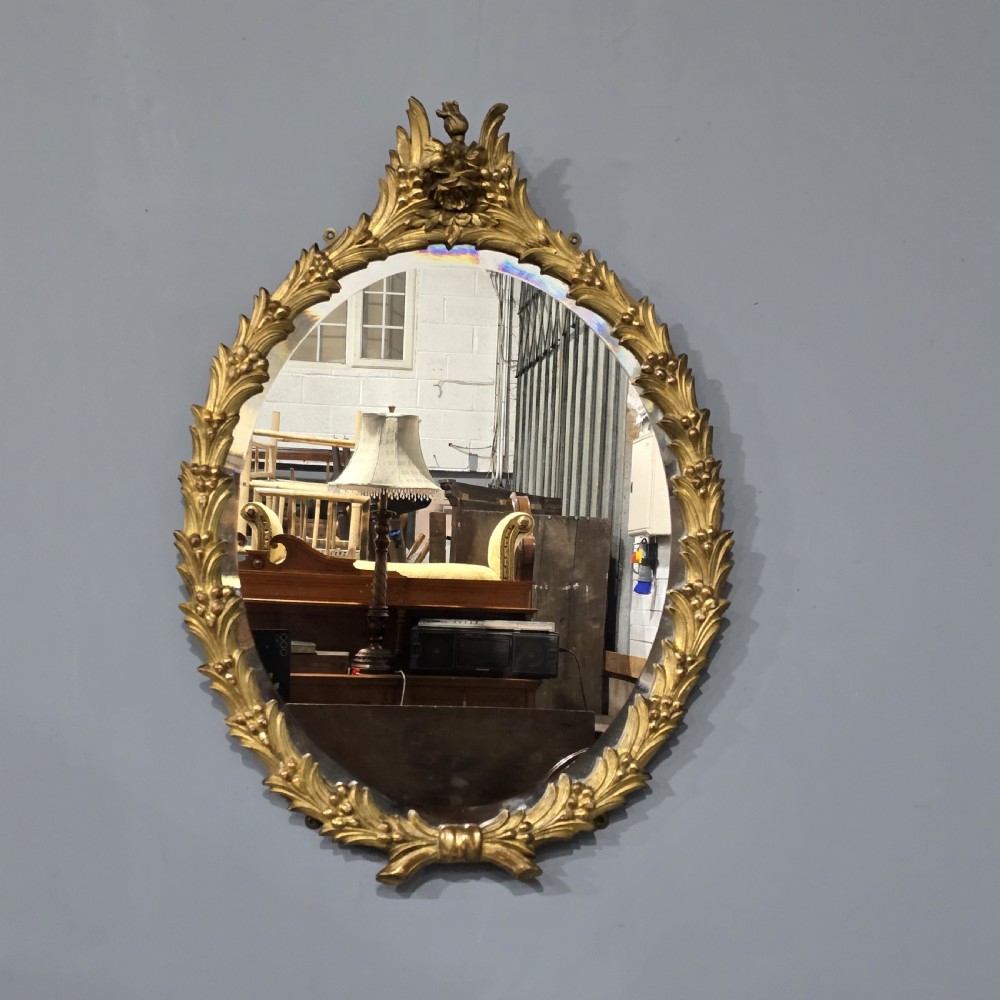 An oval wall mirror featuring a gilded carved wood frame adorned with acanthus leaves and a floral crest top motif. View II