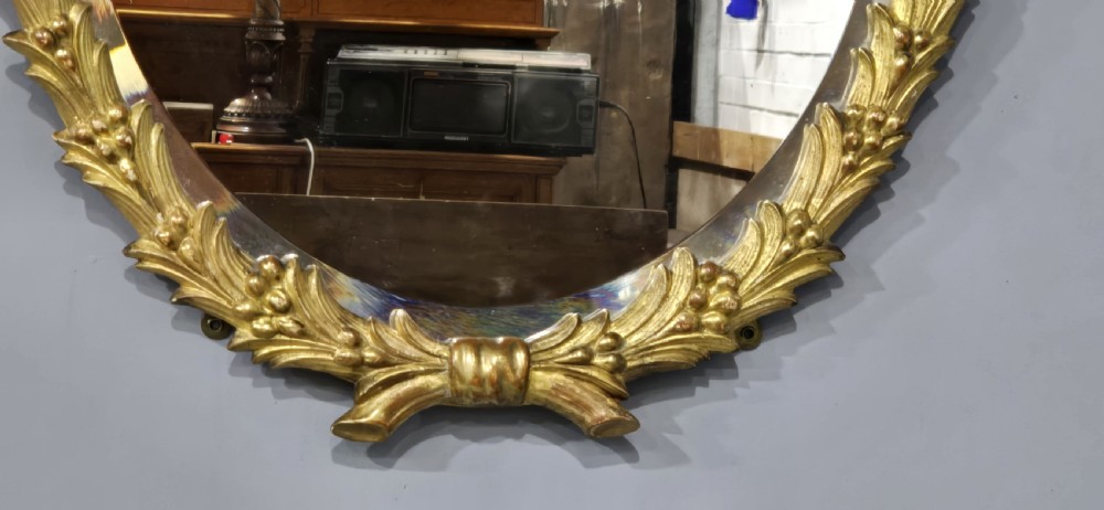 An oval wall mirror featuring a gilded carved wood frame adorned with acanthus leaves and a floral crest top motif. View IV