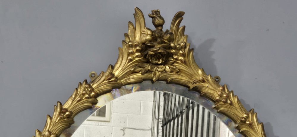 An oval wall mirror featuring a gilded carved wood frame adorned with acanthus leaves and a floral crest top motif. View V