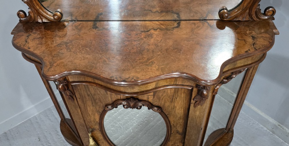 An antique burr walnut side cabinet featuring an arched top mirror with a carved cresting and glass mirrored door panels. View VI