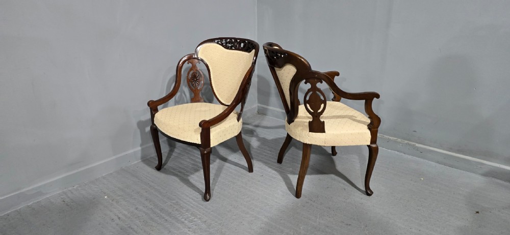 A pair of antique Victorian mahogany elbow chairs featuring carved crest rails, pierced arm supports, cabriole legs, and cream upholstered seats. View X