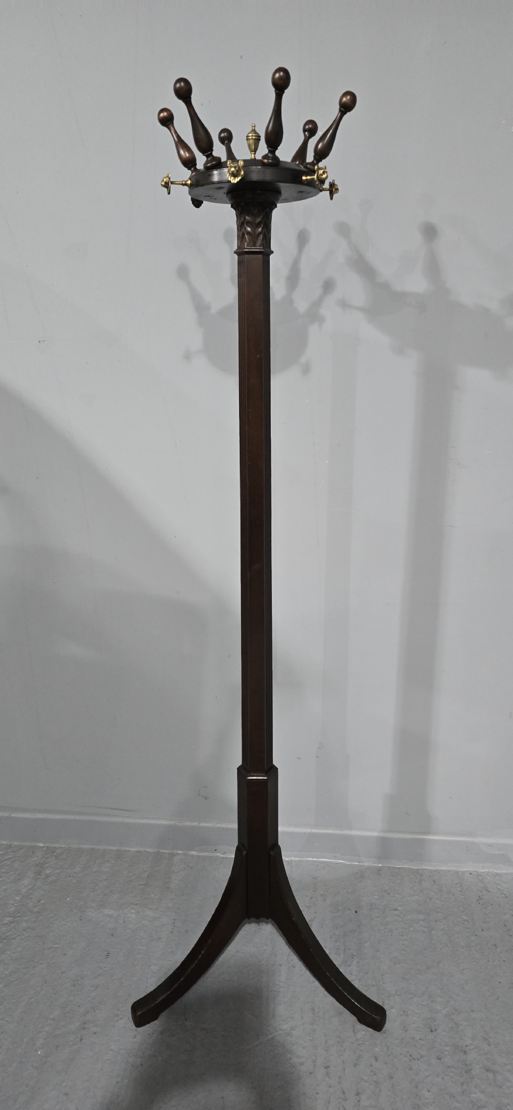 A tall dark mahogany freestanding coat stand featuring turned wooden pegs, brass accents, a fluted column, and a curved tripod base.