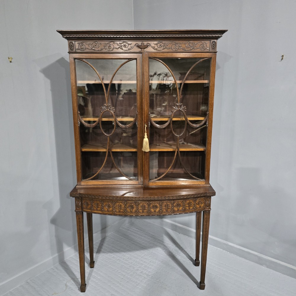An Adam style mahogany bookcase on stand featuring carved urn frieze ornamentation, astragal glazed doors, and slender tapered legs.