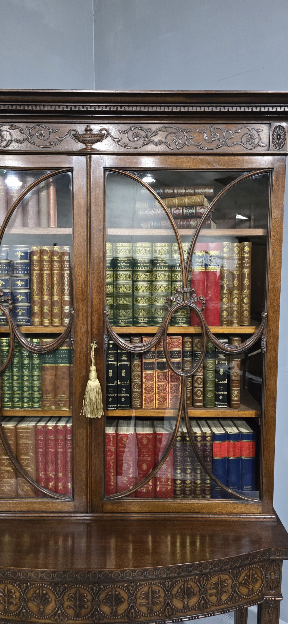 An Adam style mahogany bookcase on stand featuring carved urn frieze ornamentation, astragal glazed doors, and slender tapered legs. View V