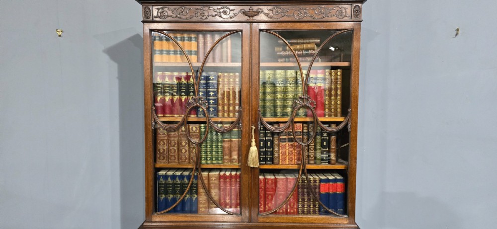 An Adam style mahogany bookcase on stand featuring carved urn frieze ornamentation, astragal glazed doors, and slender tapered legs. View VI