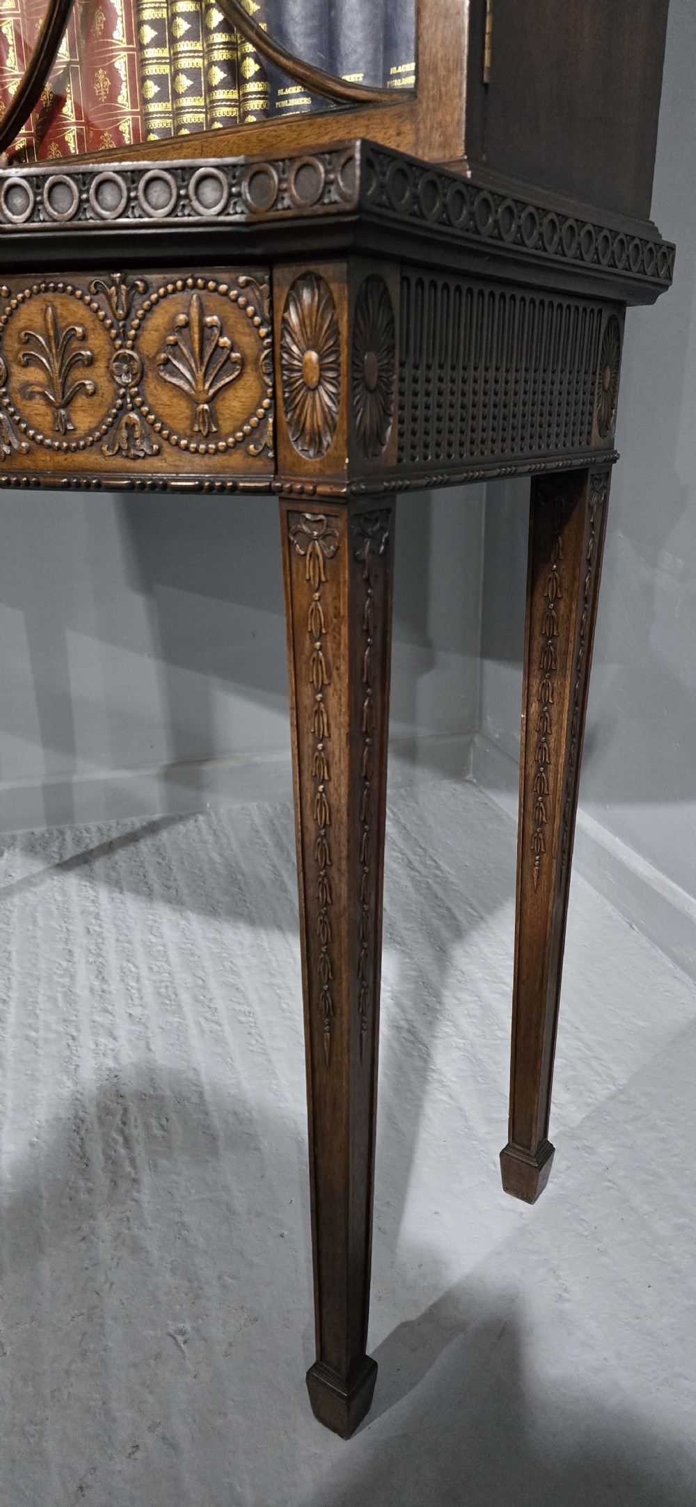 An Adam style mahogany bookcase on stand featuring carved urn frieze ornamentation, astragal glazed doors, and slender tapered legs. View XI
