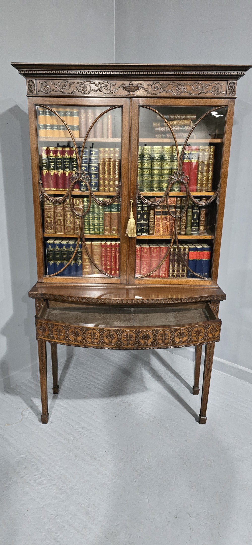 An Adam style mahogany bookcase on stand featuring carved urn frieze ornamentation, astragal glazed doors, and slender tapered legs. View XII