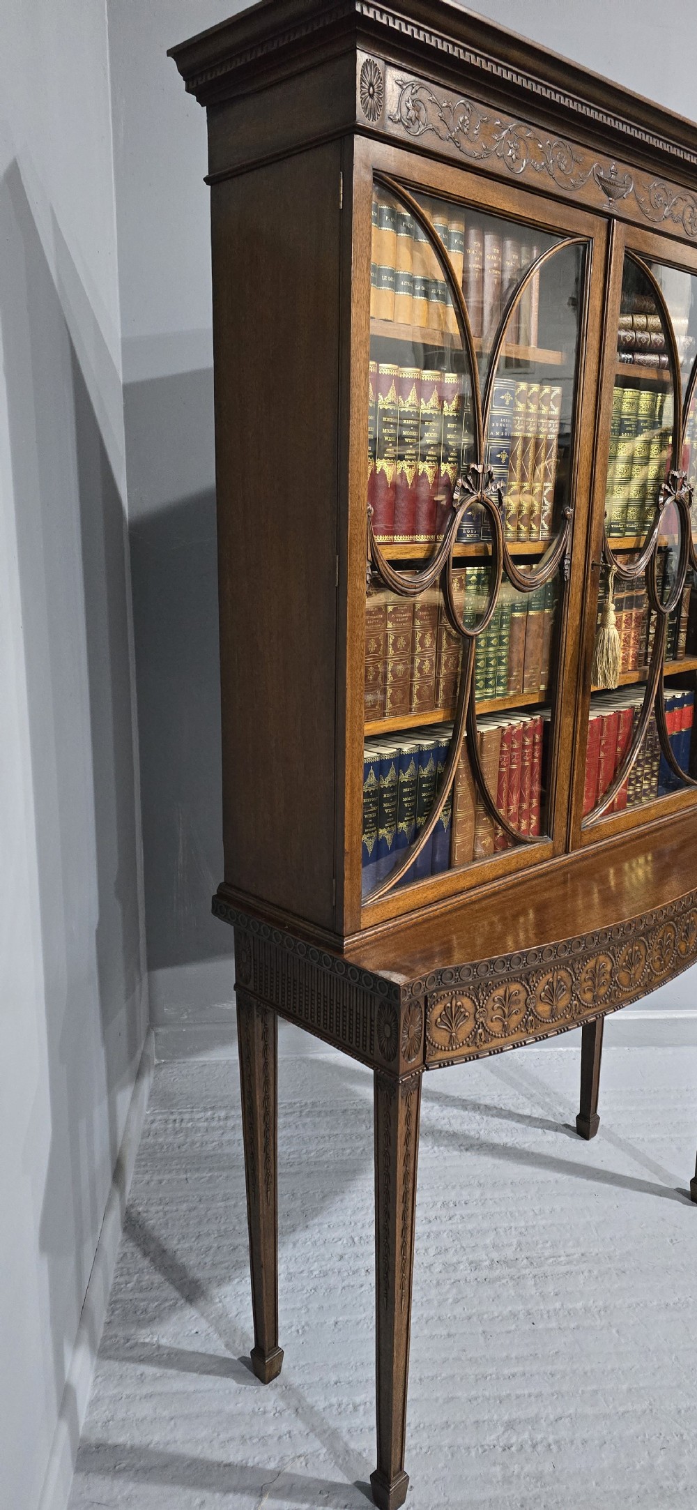 An Adam style mahogany bookcase on stand featuring carved urn frieze ornamentation, astragal glazed doors, and slender tapered legs. View XV