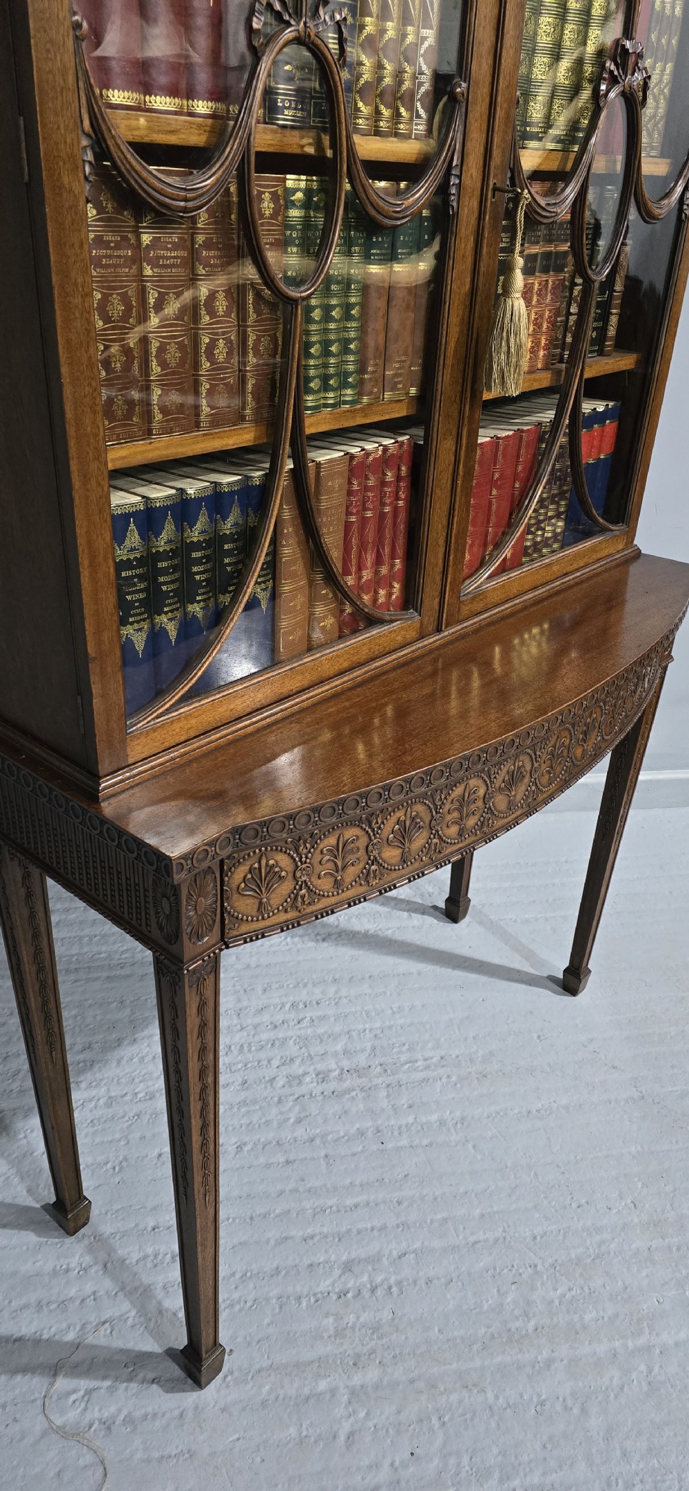 An Adam style mahogany bookcase on stand featuring carved urn frieze ornamentation, astragal glazed doors, and slender tapered legs. View XVI