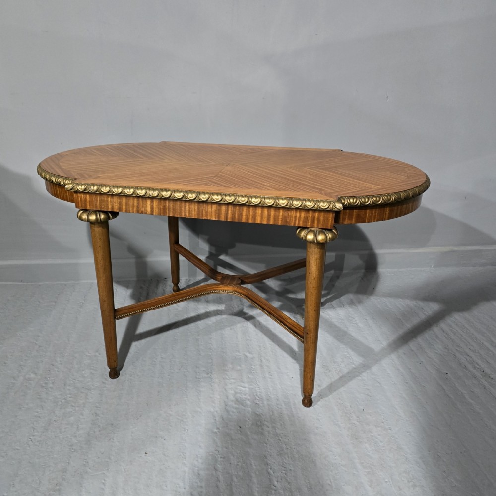 An oval satinwood coffee table featuring a parquetry top, gilt egg-and-dart rim moulding, fluted tapered legs, and an X-frame stretcher.