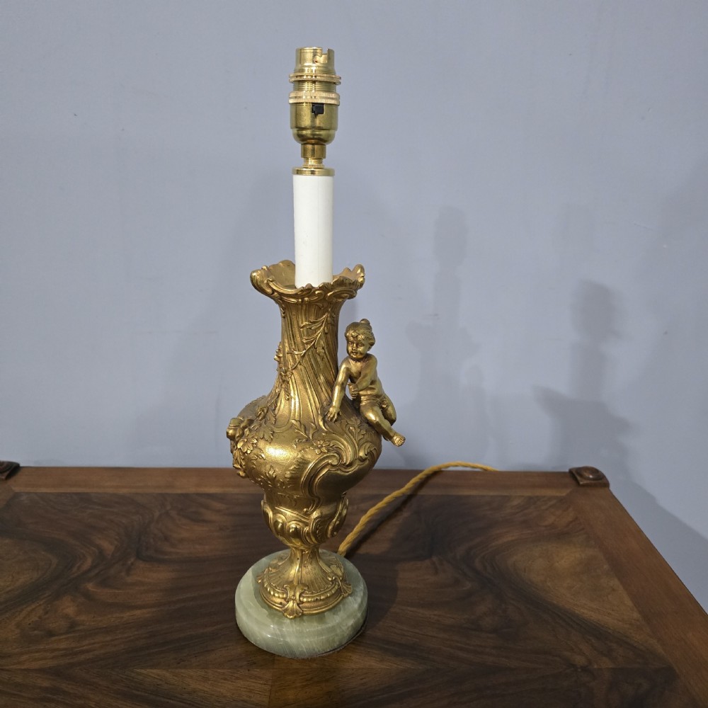 A gilt bronze table lamp featuring a cherub figure seated on a foliage-embossed urn base set upon a circular green onyx plinth.