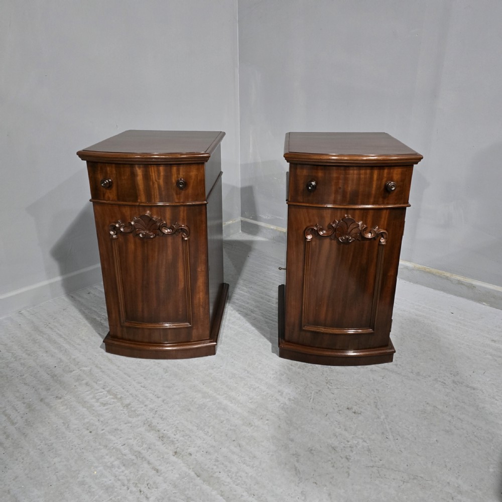 A matching pair of dark polished mahogany bedside cabinets featuring curved bow fronts, single drawers over cupboard doors, and carved decorative crestings.