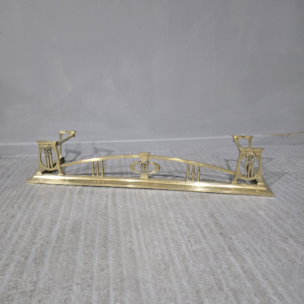 An Art Nouveau brass fireplace fender featuring a central raised arch, stylized foliate accents, and corner pedestal supports.
