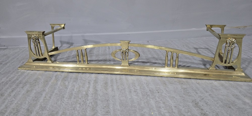 An Art Nouveau brass fireplace fender featuring a central raised arch, stylized foliate accents, and corner pedestal supports. View II