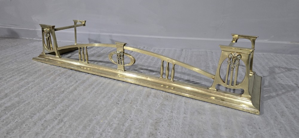 An Art Nouveau brass fireplace fender featuring a central raised arch, stylized foliate accents, and corner pedestal supports. View IV