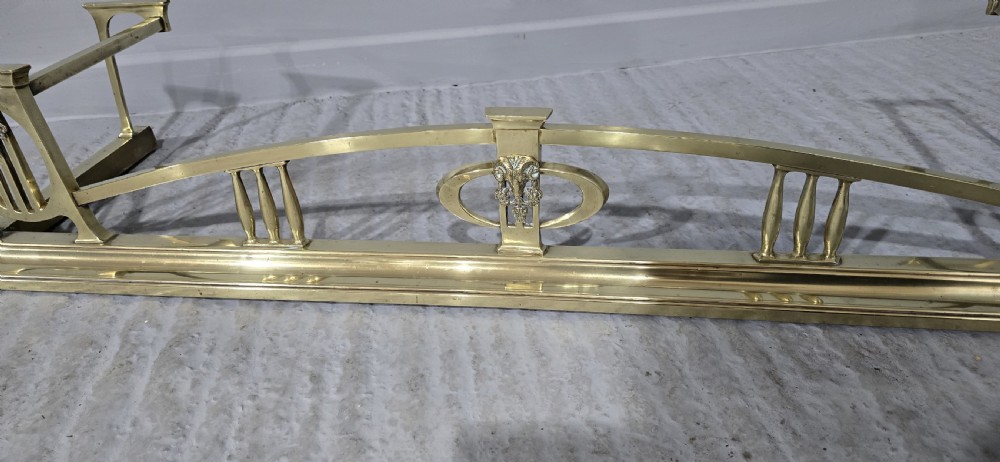 An Art Nouveau brass fireplace fender featuring a central raised arch, stylized foliate accents, and corner pedestal supports. View VI