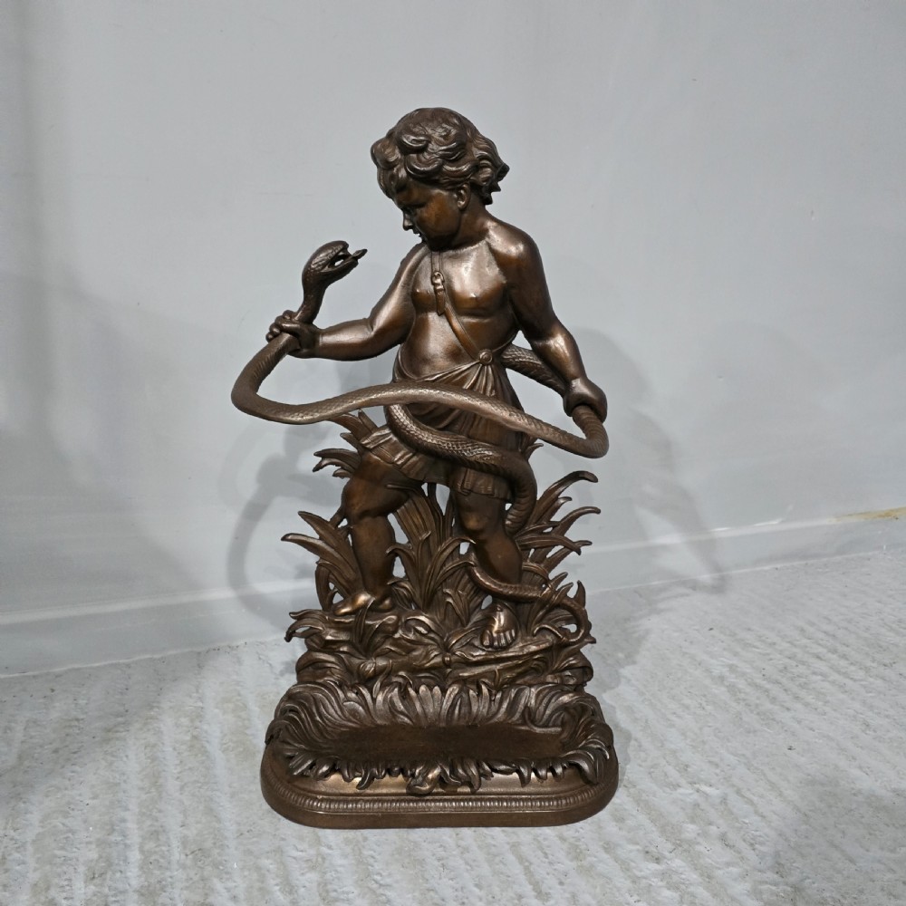 A dark bronzed cast iron stick stand featuring a sculpted figure of a child holding a coiled serpent on a leaf-patterned drip base.