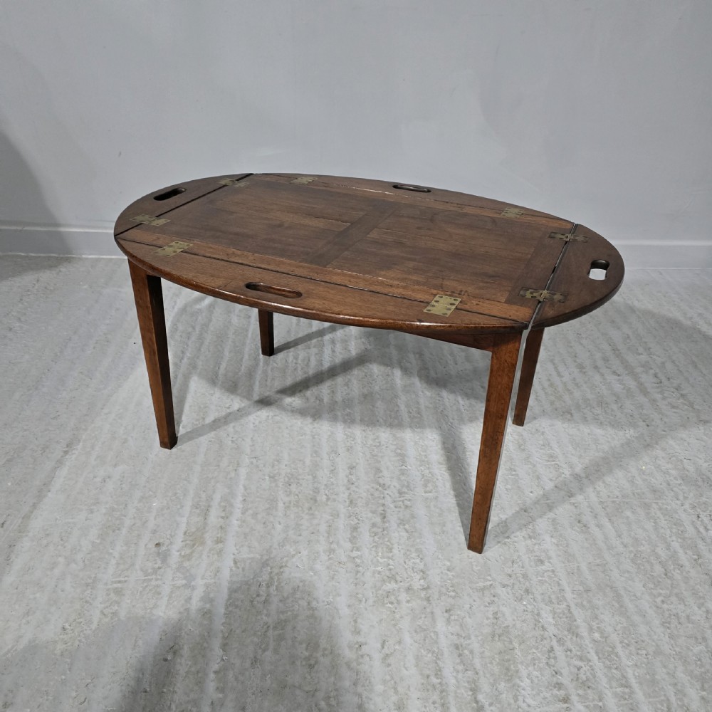 An oval Georgian mahogany butlers tray table featuring folding hinged sides with cutout handles on four straight legs.