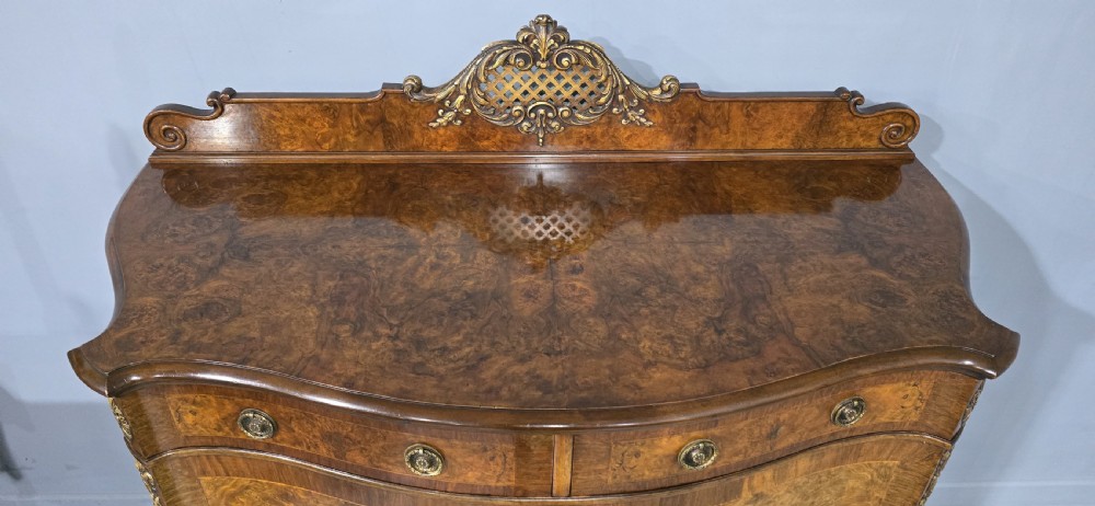 A Victorian burr walnut side cabinet featuring a serpentine front, carved upstand with pierced metal fretwork, ormolu mounts, and cabriole legs. View V
