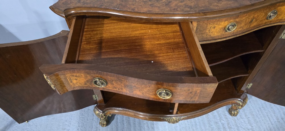 A Victorian burr walnut side cabinet featuring a serpentine front, carved upstand with pierced metal fretwork, ormolu mounts, and cabriole legs. View IX
