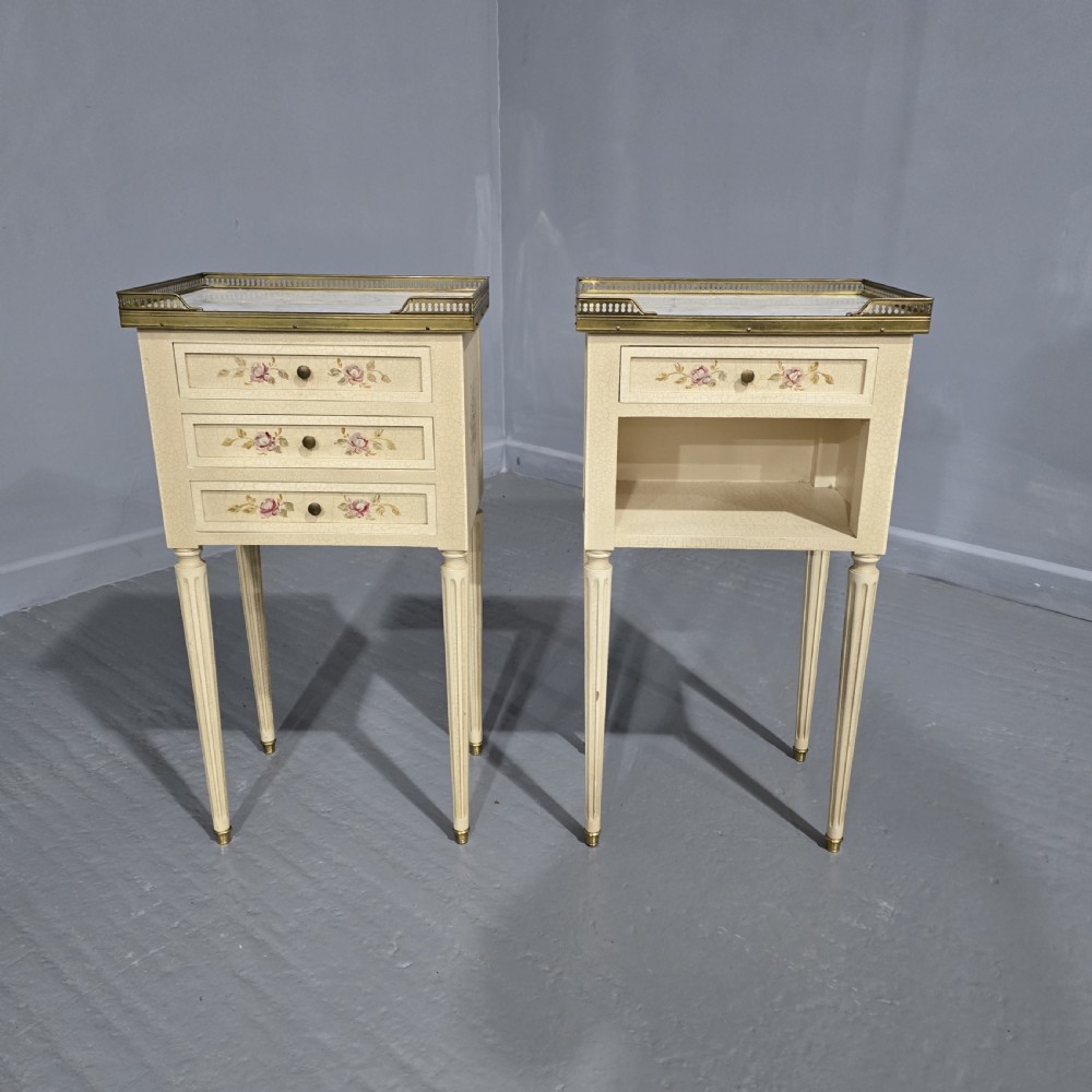 A pair of cream painted French bedside tables featuring brass gallery edges, marble inset tops, hand-painted floral drawer details, and slender fluted tapered legs.