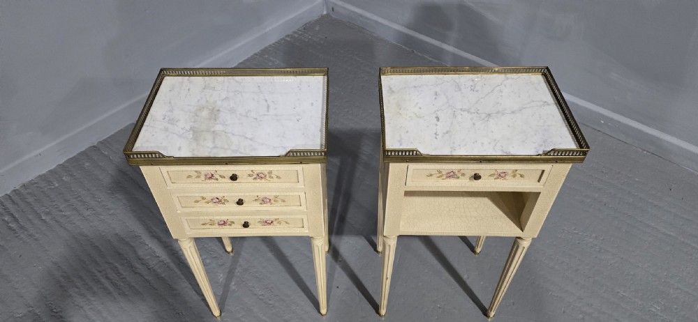A pair of cream painted French bedside tables featuring brass gallery edges, marble inset tops, hand-painted floral drawer details, and slender fluted tapered legs. View II