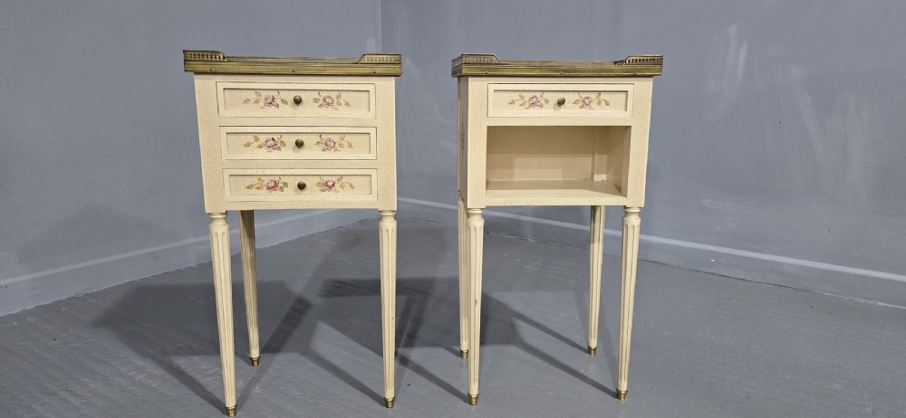 A pair of cream painted French bedside tables featuring brass gallery edges, marble inset tops, hand-painted floral drawer details, and slender fluted tapered legs. View VI