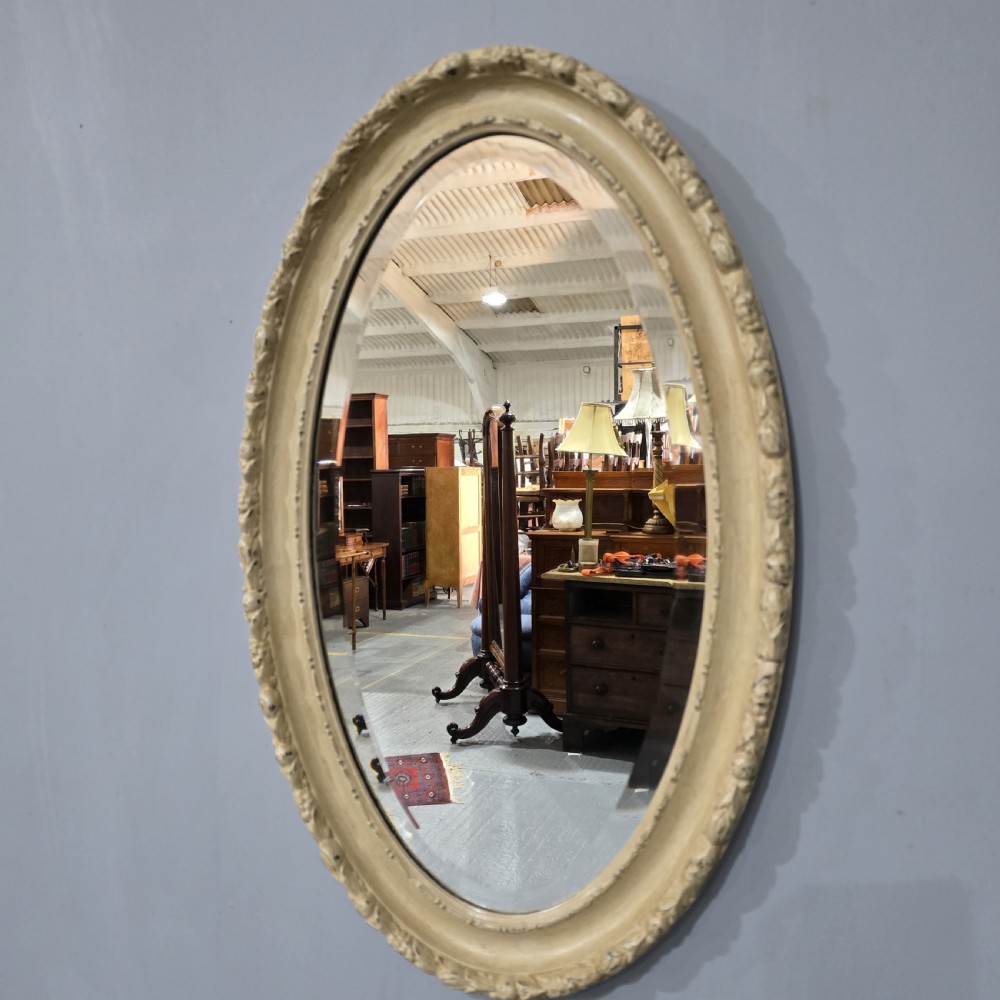 An oval Victorian wall mirror featuring a painted off-white frame with moulded foliage details around the border.