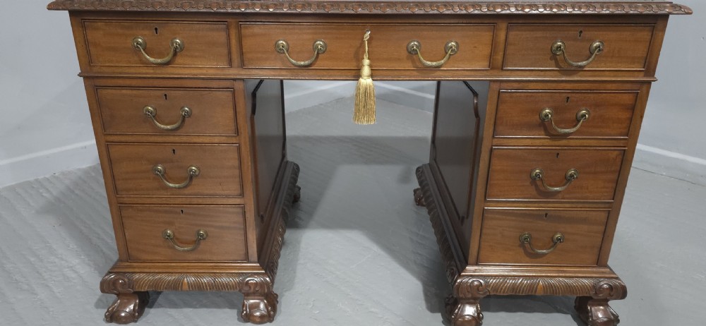 An antique mahogany pedestal desk featuring a dark leather inset surface, brass swan-neck handles, eight side drawers, a central frieze drawer, and carved claw feet. View II