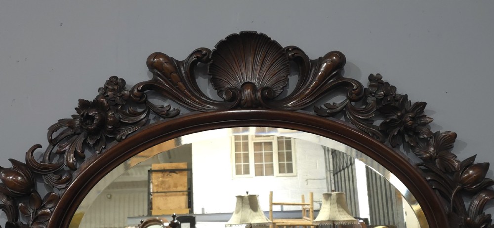 A dark mahogany oval wall mirror featuring deeply hand-carved floral motifs, a shell crest at the top, and a bevelled mirror plate. View III