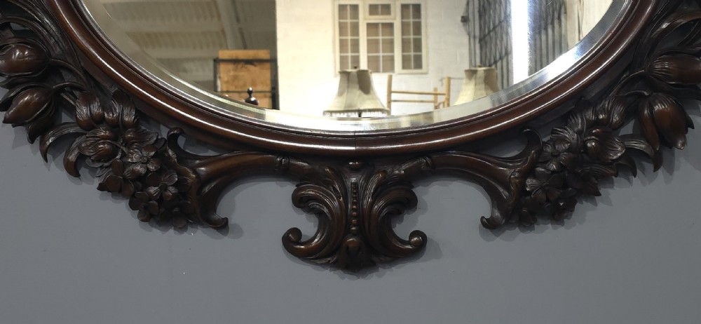A dark mahogany oval wall mirror featuring deeply hand-carved floral motifs, a shell crest at the top, and a bevelled mirror plate. View IV