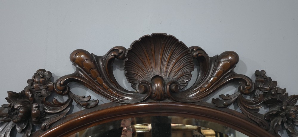 A dark mahogany oval wall mirror featuring deeply hand-carved floral motifs, a shell crest at the top, and a bevelled mirror plate. View VII