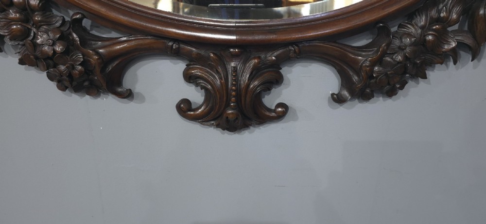 A dark mahogany oval wall mirror featuring deeply hand-carved floral motifs, a shell crest at the top, and a bevelled mirror plate. View IX