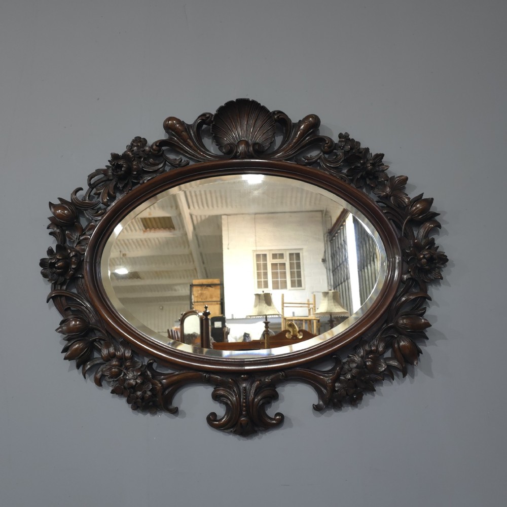 A dark mahogany oval wall mirror featuring deeply hand-carved floral motifs, a shell crest at the top, and a bevelled mirror plate.
