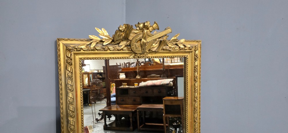 A tall rectangular French gilt mirror featuring a crest with carved musical instruments and floral frame mouldings. View V