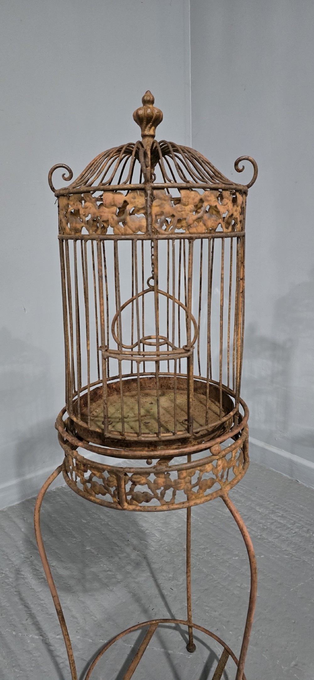 A Victorian cast iron birdcage featuring a domed roof and decorative filigree band, supported on a tall slender metal floor stand with an oxidised rust finish. View II