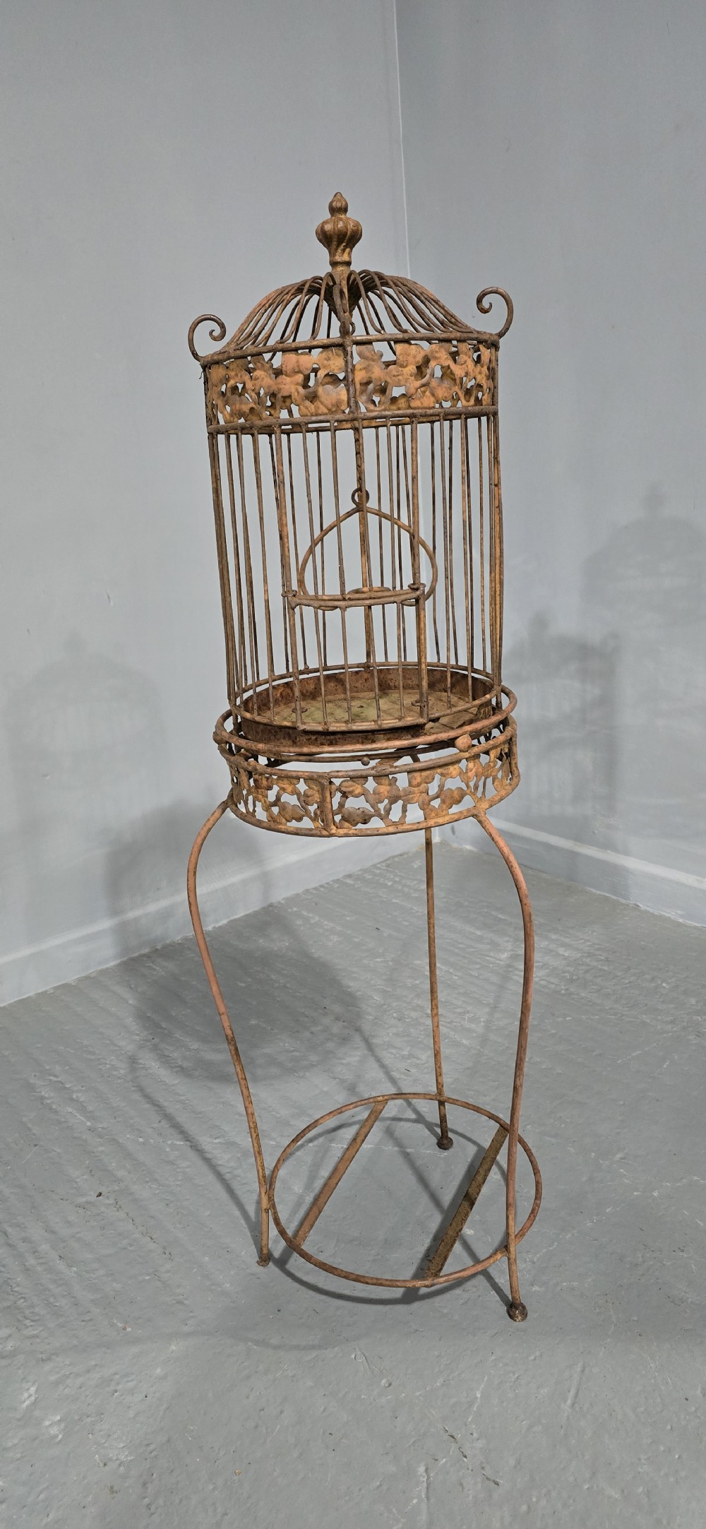 A Victorian cast iron birdcage featuring a domed roof and decorative filigree band, supported on a tall slender metal floor stand with an oxidised rust finish. View III