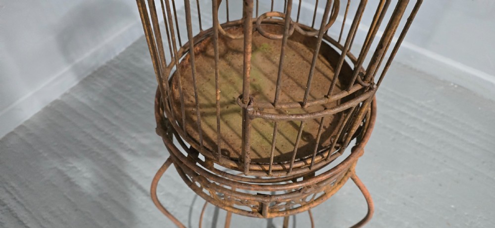 A Victorian cast iron birdcage featuring a domed roof and decorative filigree band, supported on a tall slender metal floor stand with an oxidised rust finish. View VI