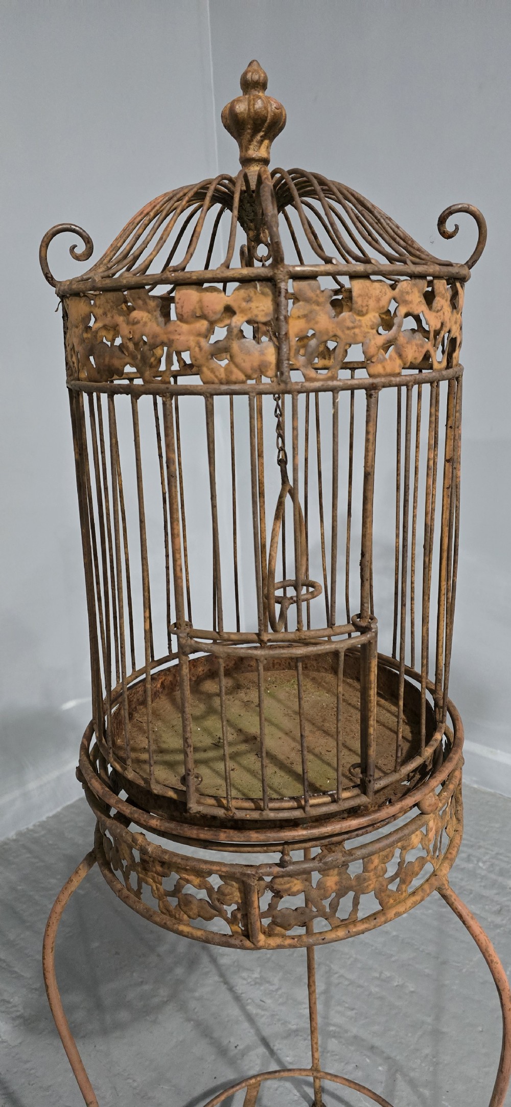 A Victorian cast iron birdcage featuring a domed roof and decorative filigree band, supported on a tall slender metal floor stand with an oxidised rust finish. View VII