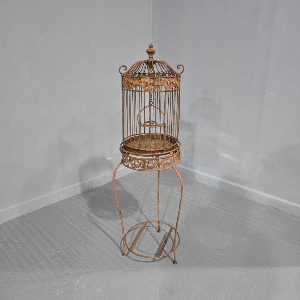 A Victorian cast iron birdcage featuring a domed roof and decorative filigree band, supported on a tall slender metal floor stand with an oxidised rust finish.