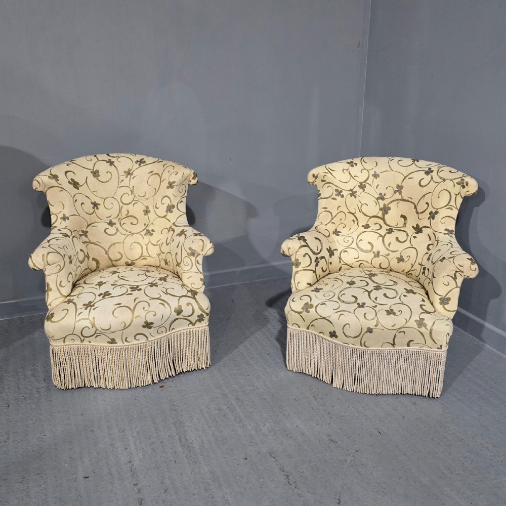 A pair of fully upholstered French Empire style armchairs covered in cream fabric with a green scroll pattern and deep tassel fringe skirting.