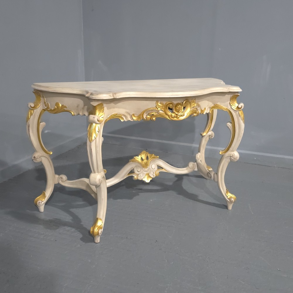 A French Rococo style console table featuring off-white painted wood, carved parcel-gilt details, cabriole legs, and a shaped stretcher base.